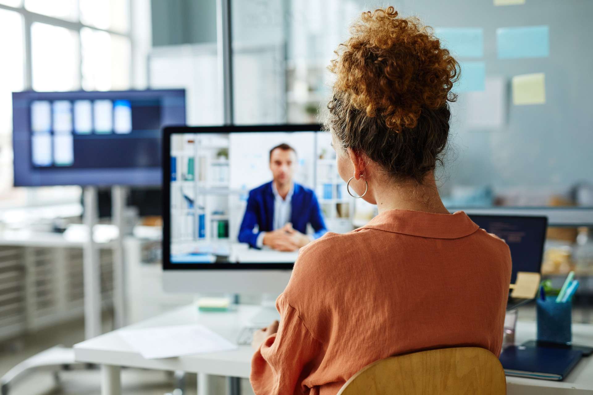  Woman participating in a virtual meeting, watching a professional man speak on a computer screen in a modern office setting.