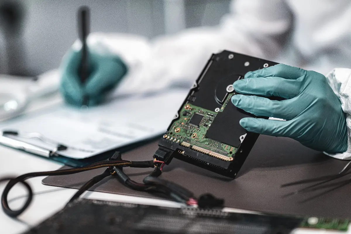 Gloved hands examining a disassembled hard drive during a digital forensics process, with notes being taken on a clipboard in the background.