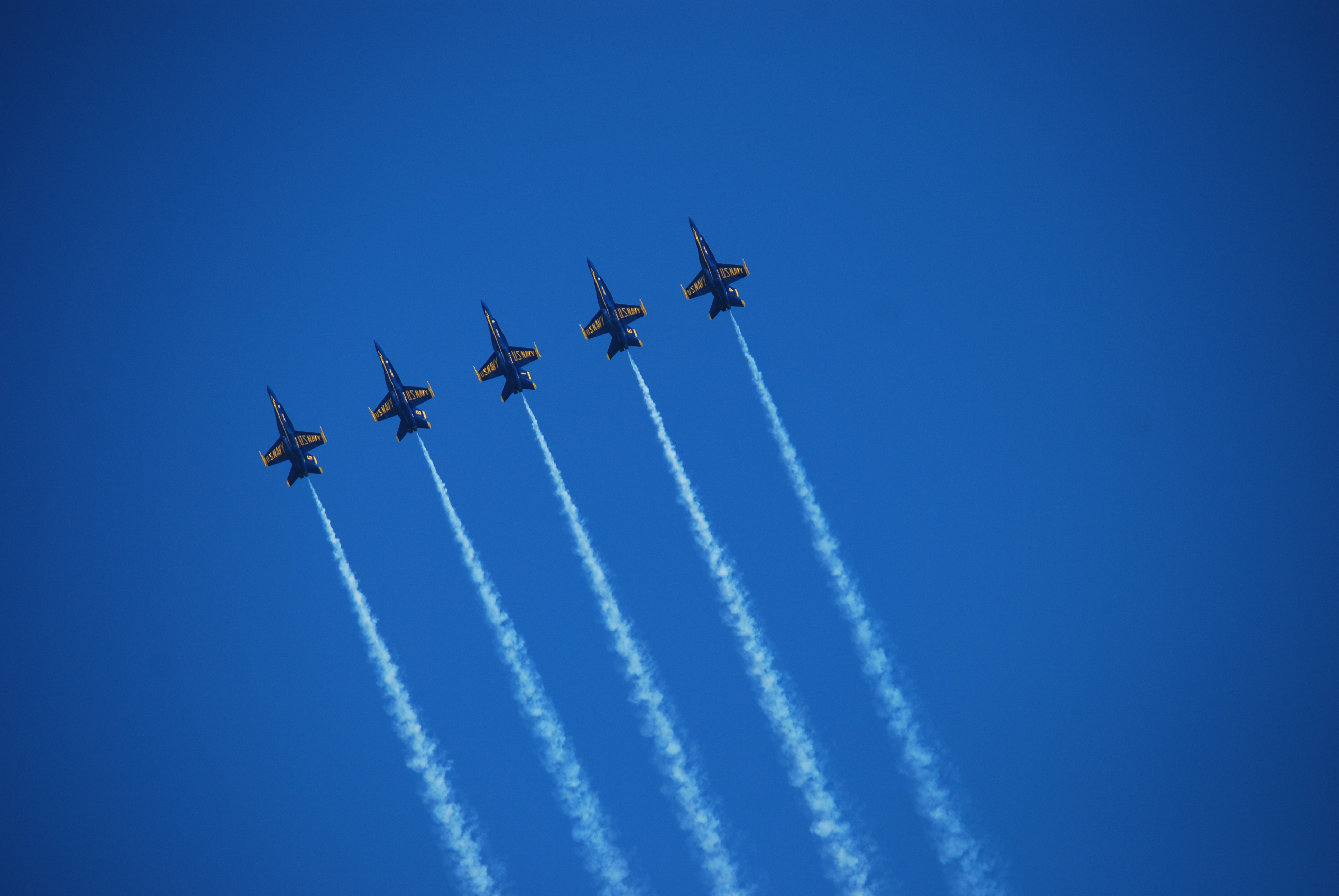 Five jet airplanes flying in a tight formation against a clear blue sky, leaving white smoke trails behind them.