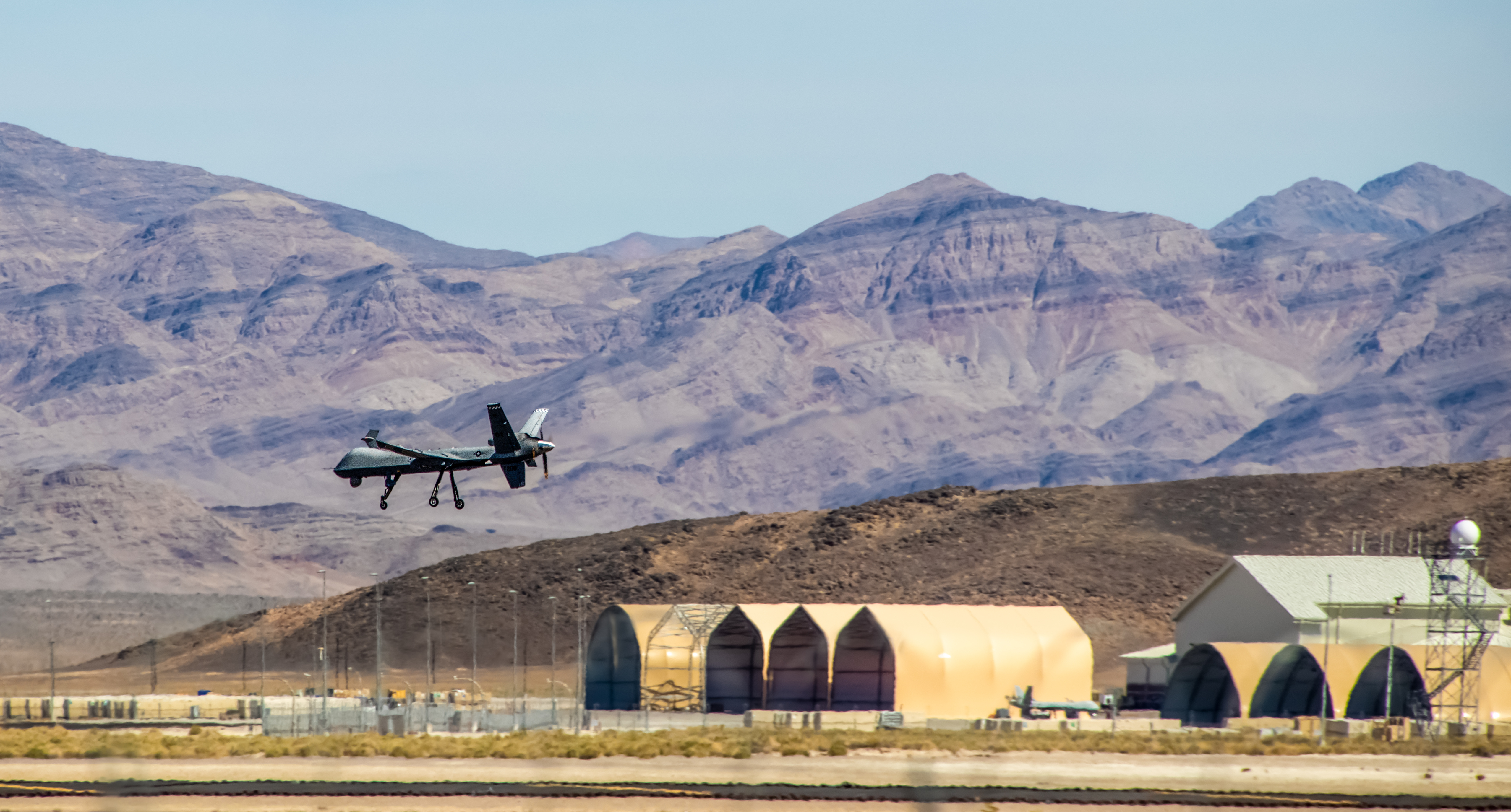 A military drone aircraft is taking off from a desert airbase with hangars and radar structures in the background, set against rugged mountain terrain.