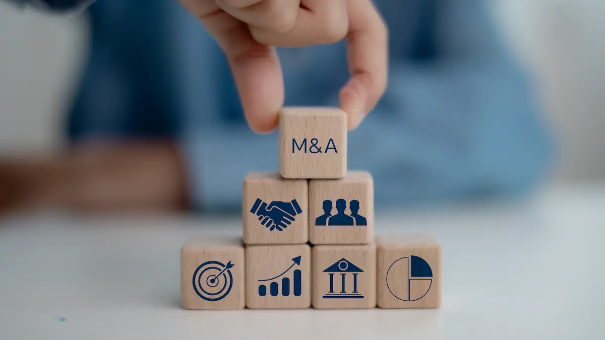The image depicts a hand placing a wooden block labeled "M&A" at the top of a pyramid-shaped stack of six other wooden blocks. The blocks are arranged in a pyramid, with the bottom row having three blocks, the middle row having two blocks, and the top single block being placed by the hand.