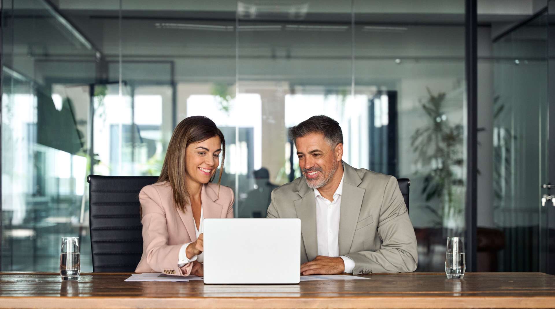 Two professionals sitting at a conference table in a modern office, smiling and reviewing information together on a laptop, symbolizing collaboration and business consulting.