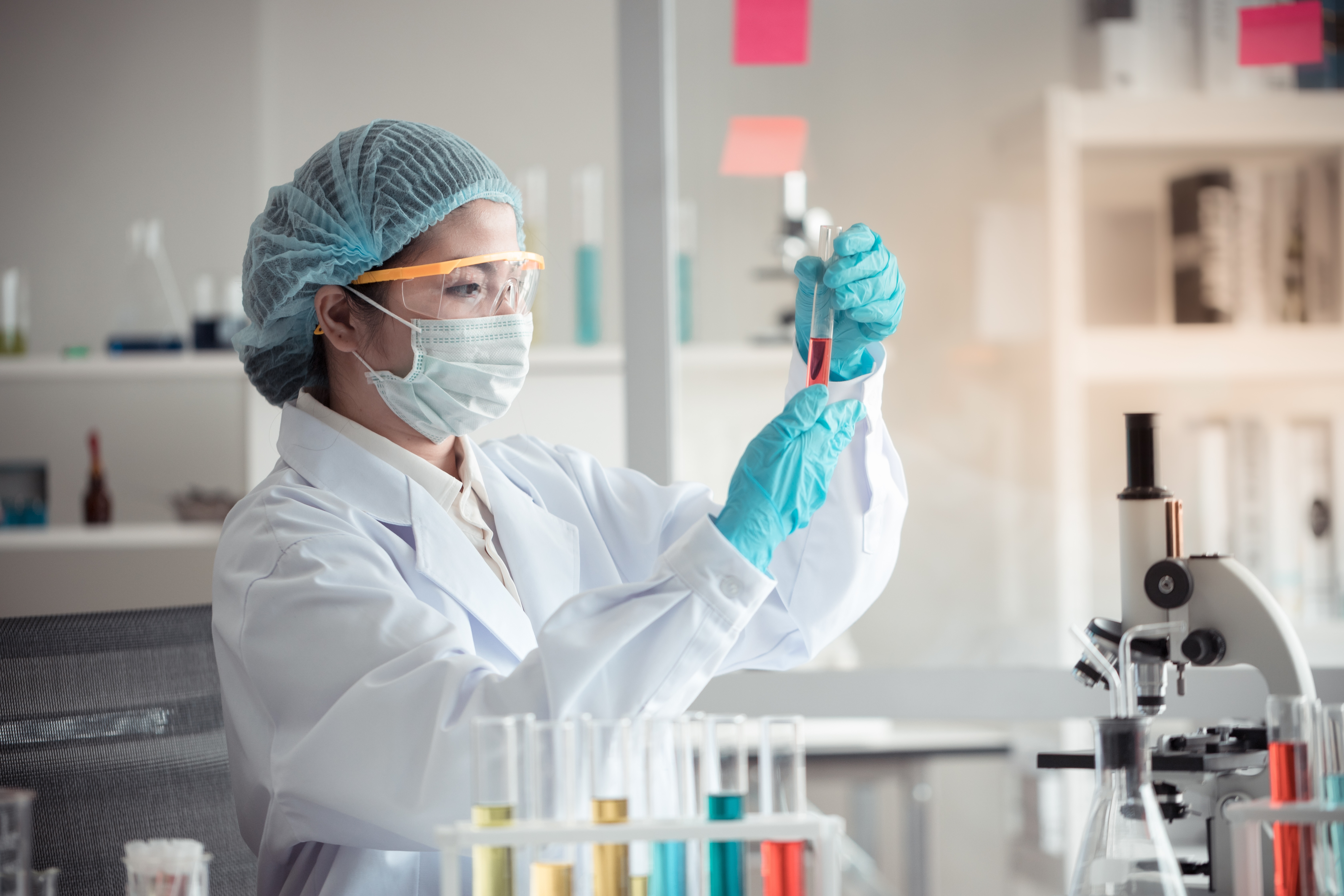 Scientist in protective gear examining a test tube with red liquid in a laboratory setting.