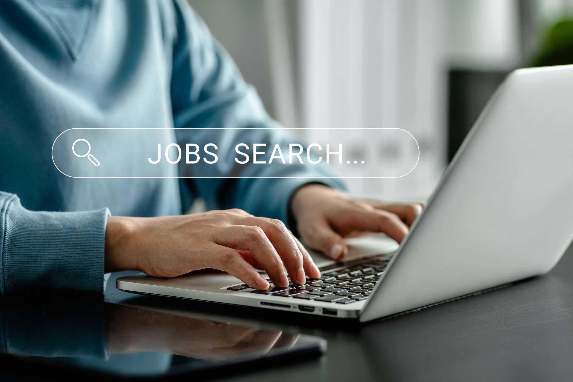  Person typing on a laptop with a digital search bar overlay displaying the words "Jobs Search," representing online job searching.